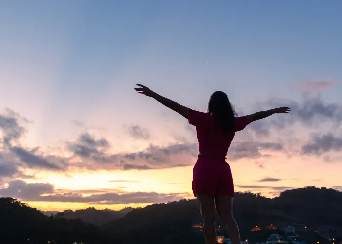 Silhouette of a young woman with arms raised and the sky after sunset in the background.