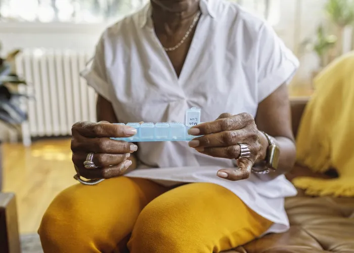 Senior black woman sits on the couch at home and takes medications from a daily pill organizer. Cropped shot does not show the woman's face.